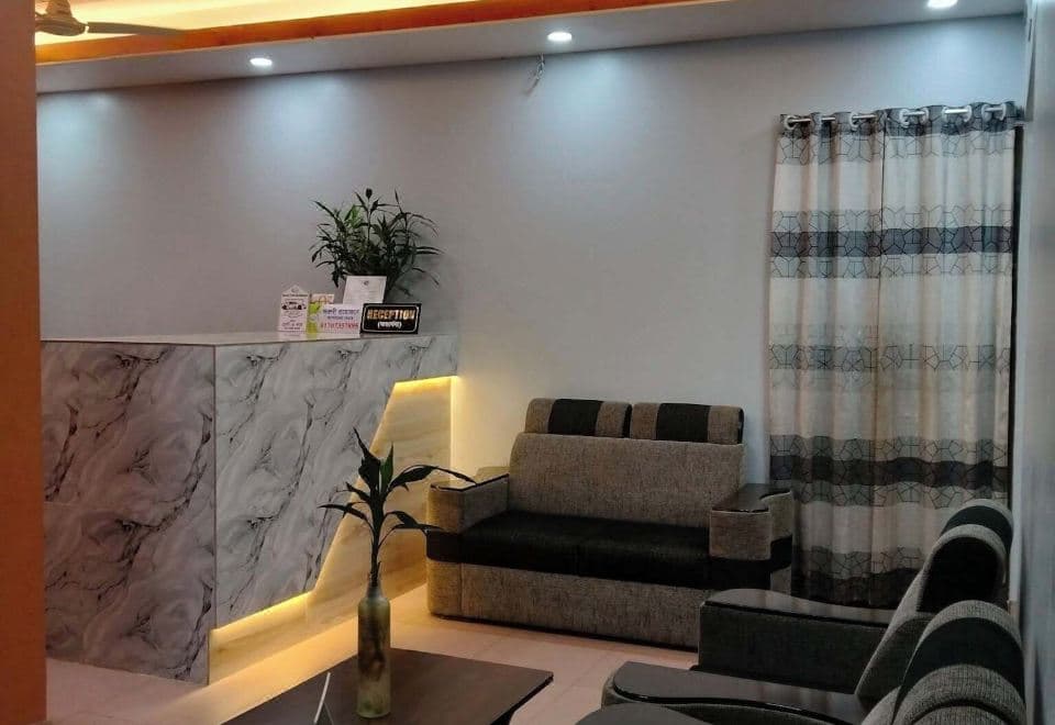 Hotel reception area with an L-shaped marble-clad front desk, a small bilingual RECEPTION sign on the counter front, potted plants, recessed downlights under a wood-slat ceiling panel, and a grey and black sofa beside a patterned floor-length curtain