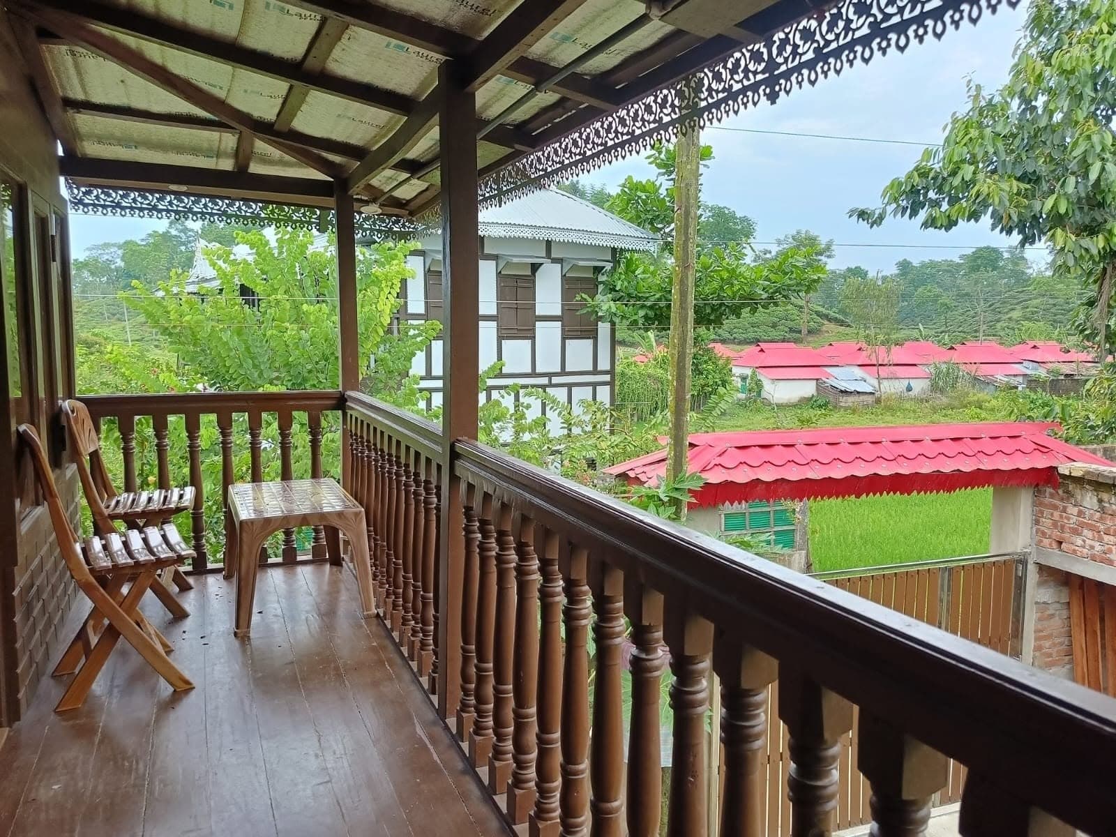 Covered wooden veranda with lathe-turned balusters and carved fretwork eaves, a folding wooden chair and plastic stool, looking out over a white cottage with dark timber framing, red tin-roofed village houses, a bright green rice plot and tea-covered hills