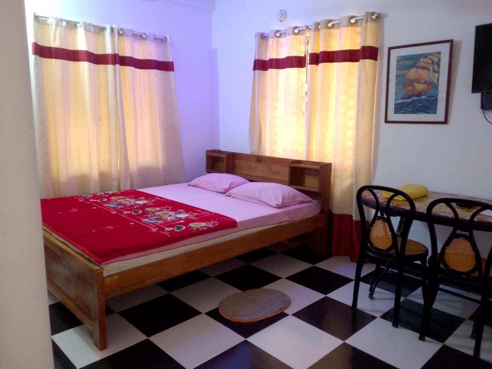 Guest bedroom with a red floral-print blanket folded across a white-sheeted wooden bed, cream curtains with a maroon band, a black-and-white checkerboard tile floor, a wall-mounted TV, a framed sailing-ship painting and a small round table with two bentwood cane-seat chairs