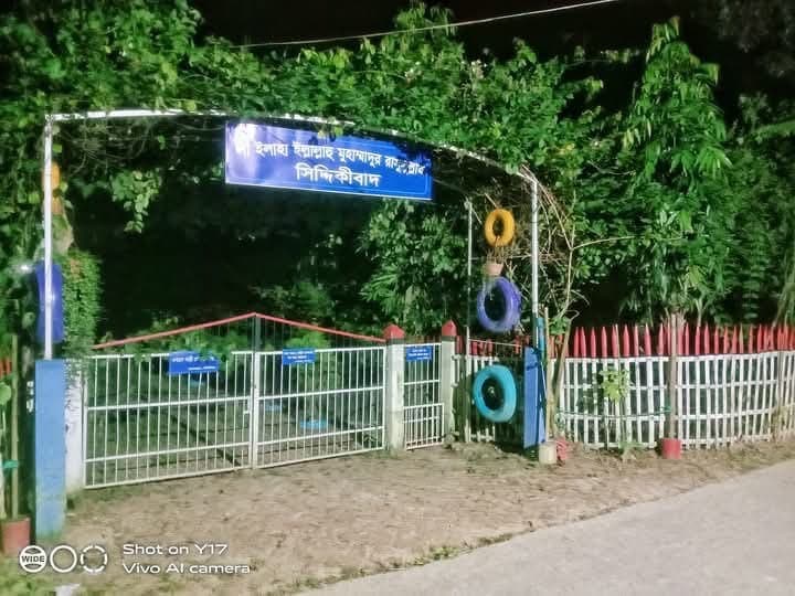 Entrance gate at night with a blue signboard reading "সিদ্দিকীবাদ" (Siddiquibad) in Bangla, flanked by a white picket fence and tyre planters