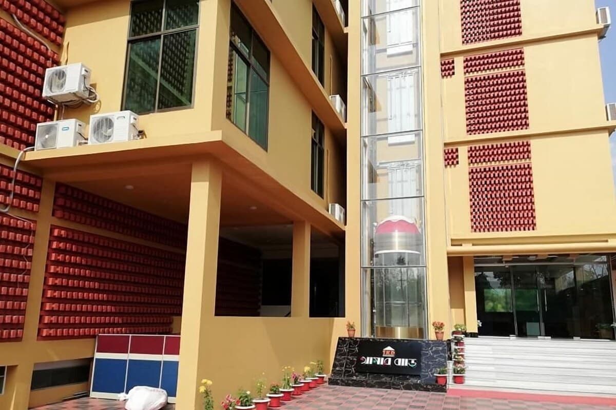 Multi-storey yellow building with red patterned wall panels, a glass elevator shaft, and the black marble SKD Amar Bari entrance sign at the base of the steps